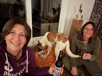 Brown and white dog stands on a blanket between two smiling women holding beer bottles indoors